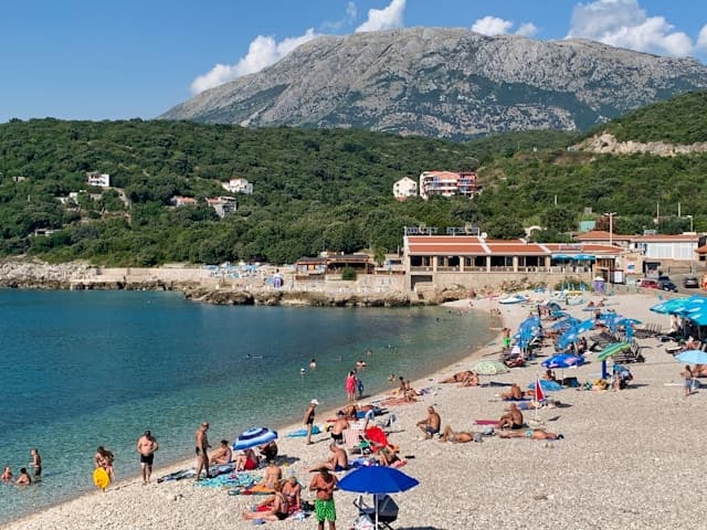 Tłoczna plaża kamienista z krystaliczną wodą i górami w tle.