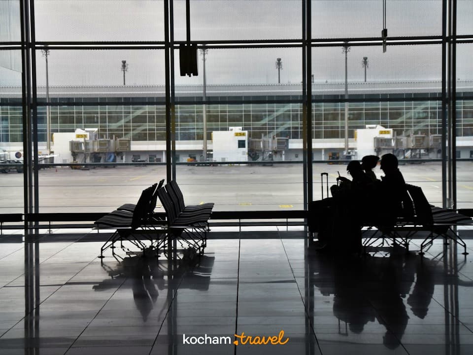 Sylwetki podróżnych czekających na lotnisku, widok na płytę postojową.
