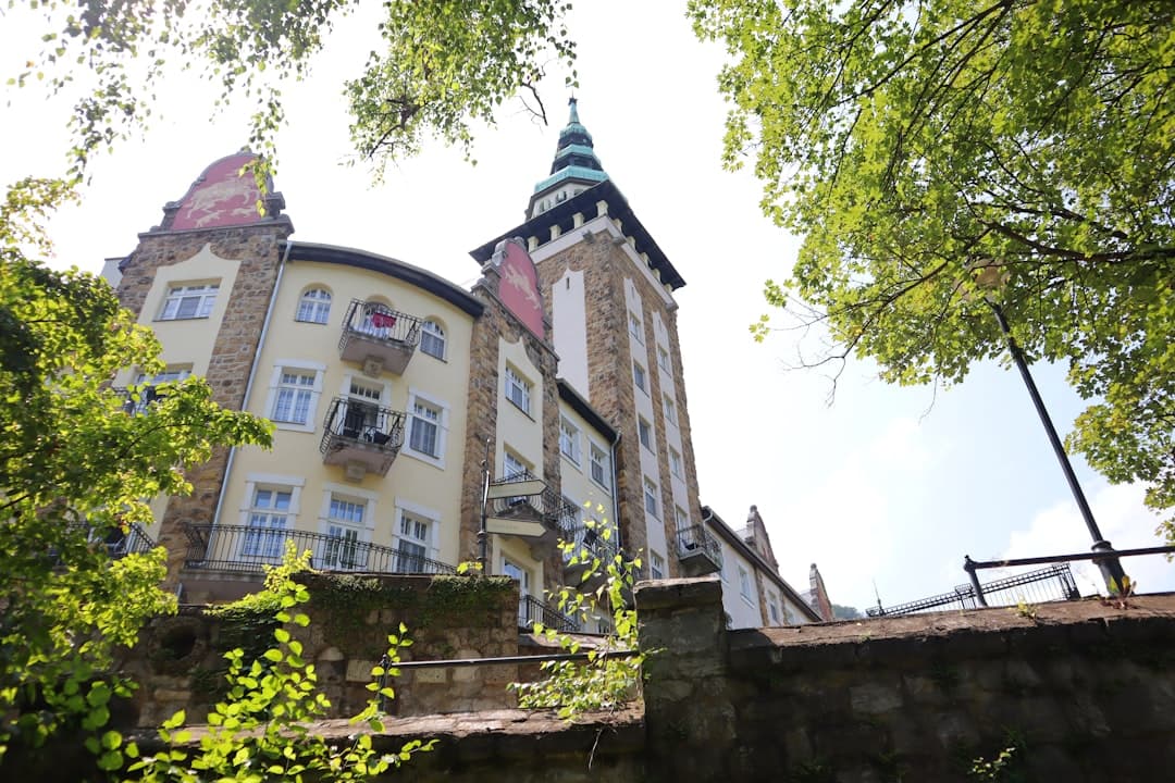 Secesyjny budynek z kamienną wieżą i zielonym dachem, otoczony zielenią.