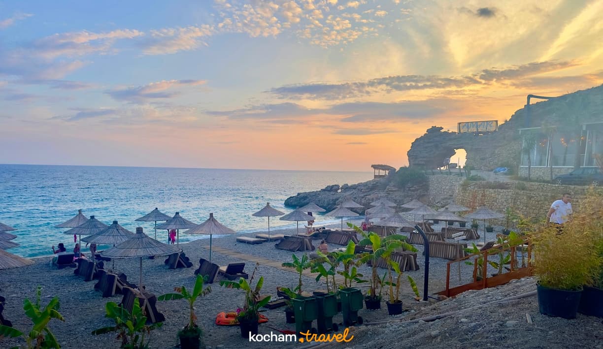 Kamienista plaża z parasolami, morzem i skalnym łukiem o zachodzie słońca.