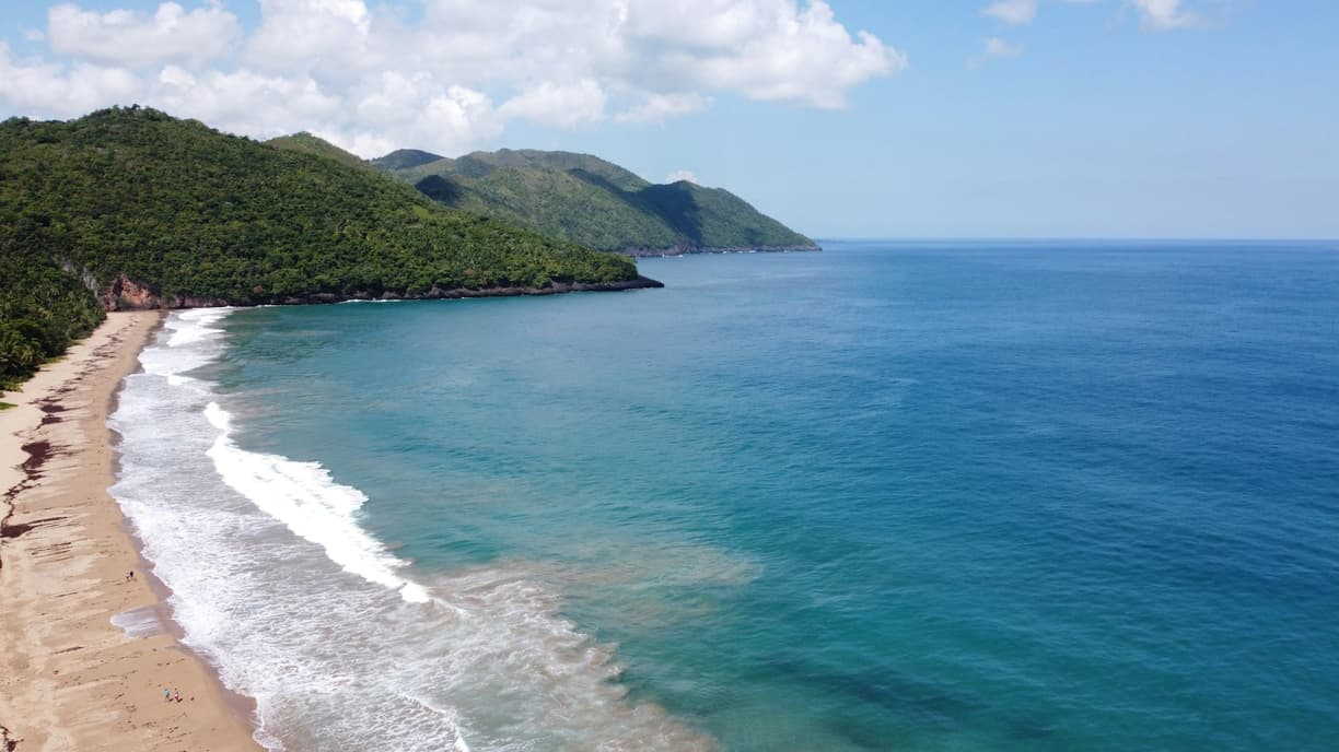 Tropikalna plaża z turkusowym oceanem, falami i zielonymi wzgórzami.