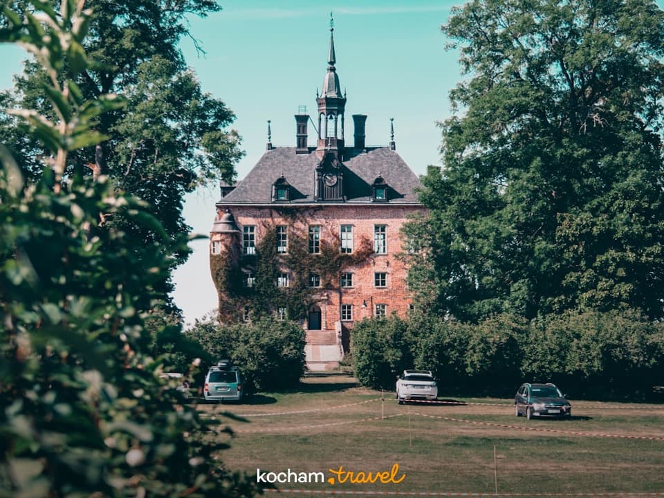Historyczny ceglany zamek z wieżą, zielone drzewa, słoneczny dzień.