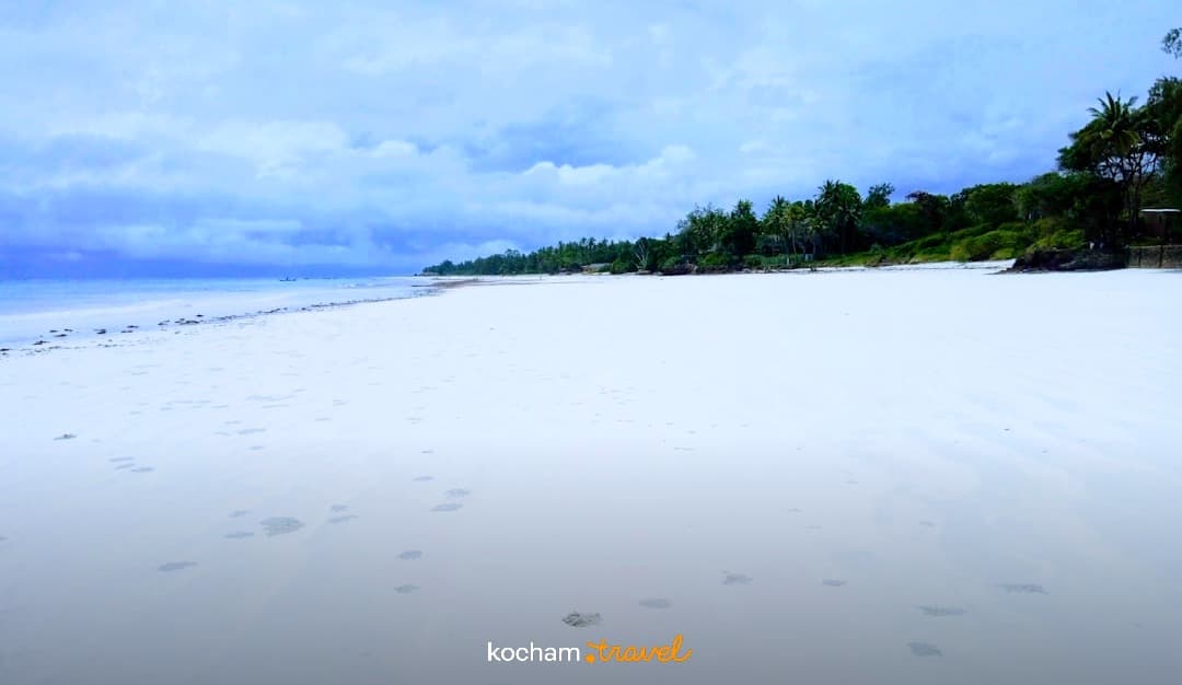 Szeroka biała plaża, błękitny ocean, palmy, pochmurne niebo.