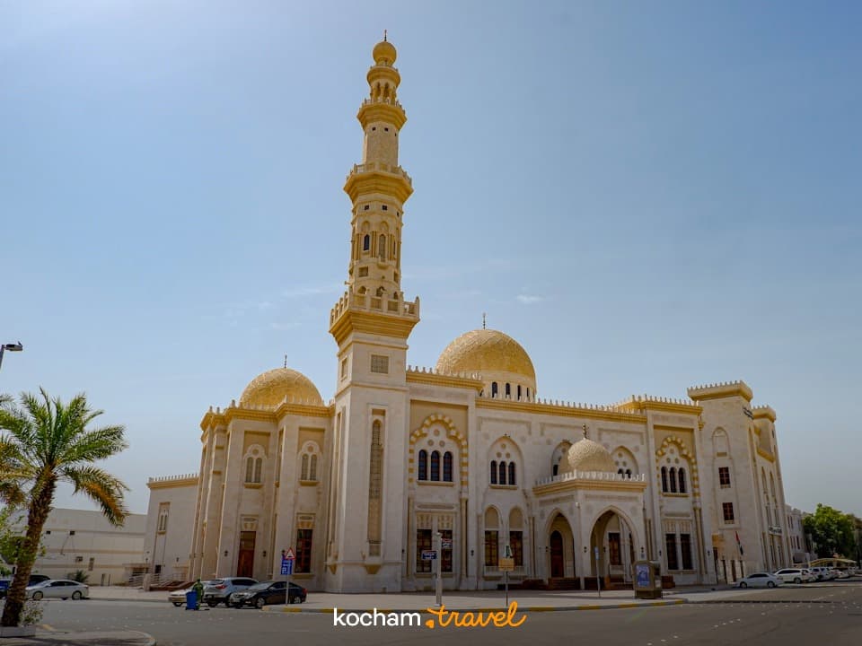 Wielki meczet z minaretem, złotymi kopułami, palmami, jasnym światłem dziennym.