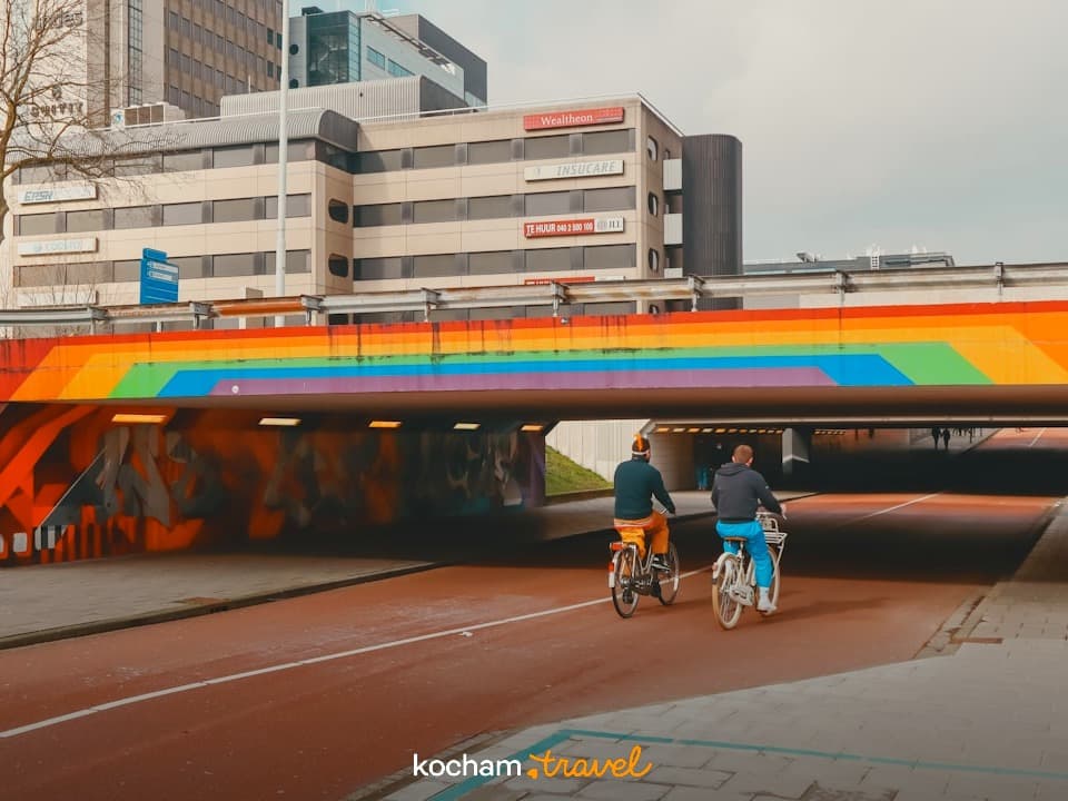 Rowerzyści przejeżdżający przez tęczowy tunel w miejskim krajobrazie.