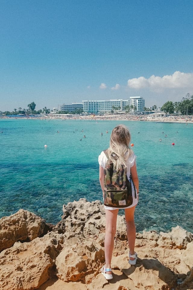 Kobieta z plecakiem na skałach podziwia turkusowe morze, plażę i hotele.